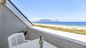 Un balcón con una mesa, una botella de vino y vistas a la playa y las montañas.