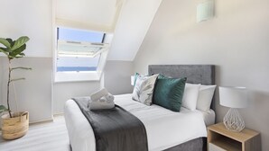 Un dormitorio con cama, almohadas, una mesita de noche con lámpara, una ventana y una planta.