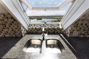 A kitchen with two stainless steel sinks, a window with a view of a lake and mountains, and checkered tile walls. - Dolphin Beach H301 (Cape Town)