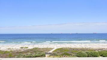 A beach with a concrete walkway leading to the shore, a clear blue sky, and three ships in the distance.
