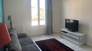 Uma sala de estar com um sofá, uma televisão sobre um móvel, uma janela com cortinas e um abajur.