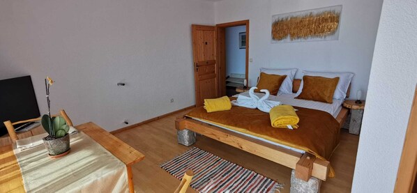 Un dormitorio con una cama de madera, una mesita con una planta en maceta y una puerta que da a otra habitación.