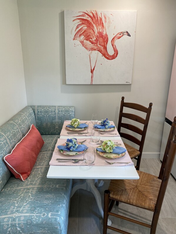 Un área de comedor con una mesa preparada para cuatro personas, un sofá azul con estampado, una silla de madera y un cuadro colgado en la pared de un flamenco.