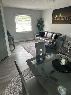 A modern living room with a glass dining table, a sofa, and a large window with blinds.