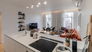 A modern kitchen with a built-in stove, dining area with a round table and chairs, a sofa, and a TV in the background. - Almada 551 - 3.1 l Luxury Living by LovelyStay (Porto)