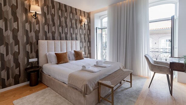 A modern hotel room with a geometric patterned wall, a large bed with white linens, a bench at the foot of the bed, a desk with a chair, and a view of the city through large windows. - Almada 551 - 3.1 l Luxury Living by LovelyStay (Porto)