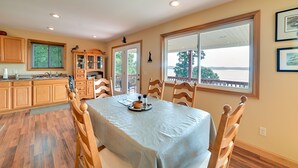 A dining area with a table set for four, wooden chairs, a kitchen with wooden cabinets, and a view through a large window. - Deck & Water View: Cozy Kentucky Lake Getaway (New Concord)