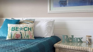 Un letto con una coperta blu, un cuscino con la scritta 'BEACH' ricamata in verde e un cartello con la scritta 'Relax' su un tavolino in vimini accanto al letto.