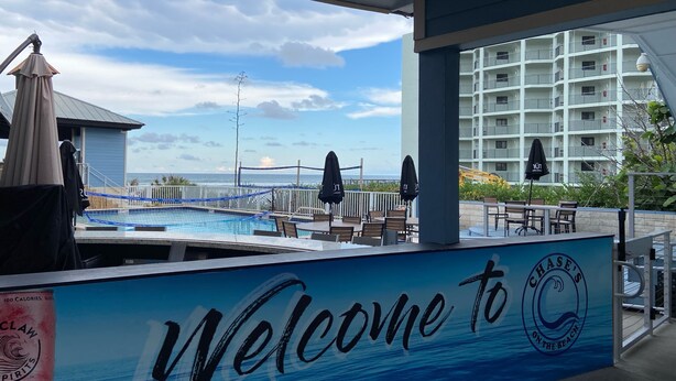 Area piscina con un cartello 'Welcome to Chase's on the Beach', posti a sedere all'aperto e vista sull'oceano e un edificio a più piani sullo sfondo.