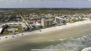 Vista aerea di una zona costiera con un hotel fronte spiaggia, un grande edificio a più piani e una spiaggia di sabbia.