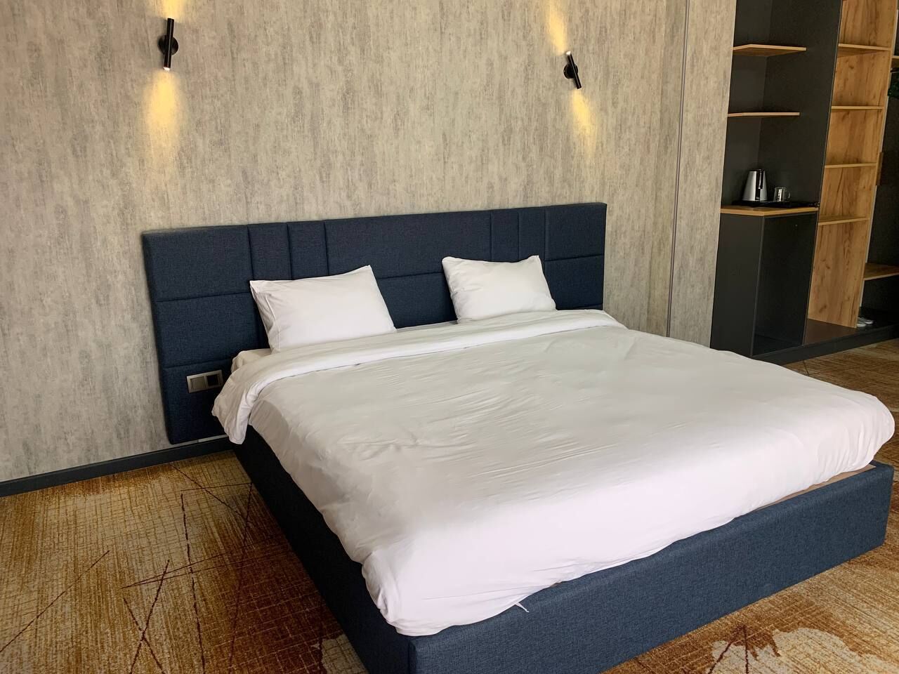 A modern hotel room with a large bed, a dark headboard, and built-in shelves with a coffee maker.