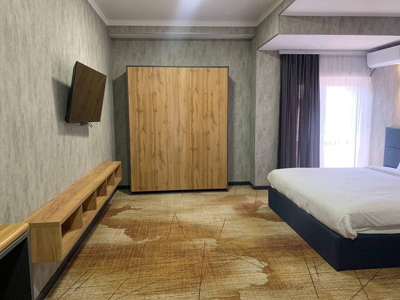 A bedroom with a bed, a wooden wardrobe, a bench, and a flat-screen TV.