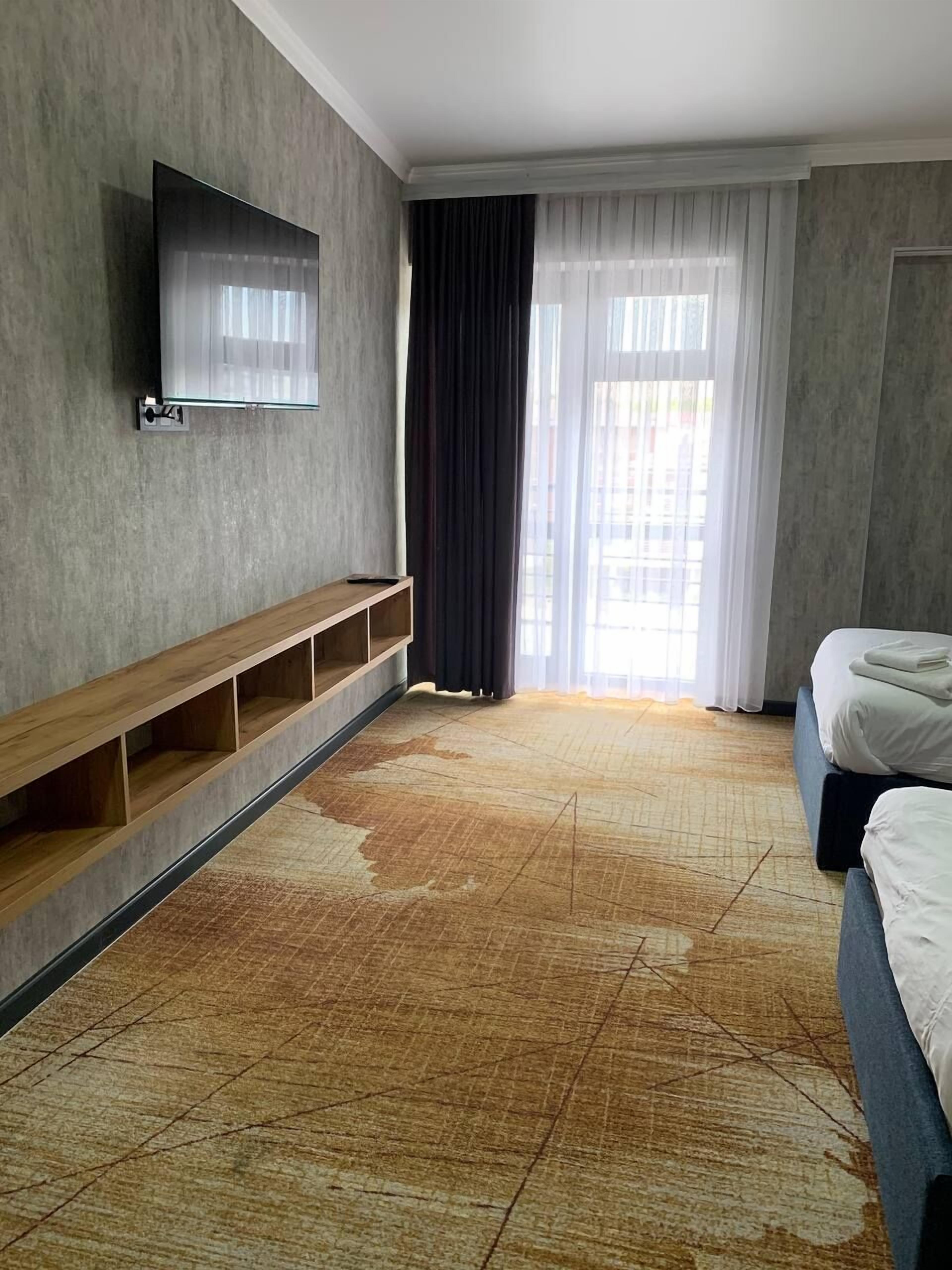 A hotel room with a large window, a flat-screen TV mounted on the wall, a wooden bench, and a bed with neatly folded towels.
