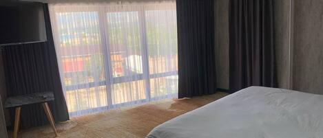 A hotel room with a large bed, a TV on a stand, and a view of a balcony with curtains.