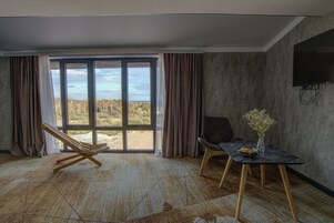 Quarto com uma grande porta de vidro deslizante que dá para uma vista de árvores e céu. Há uma cadeira de madeira, uma cadeira preta e uma pequena mesa com um vaso de flores.