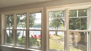 Ein Zimmer mit vier Fenstern, das auf einen See und eine Wiese mit roten Stühlen blickt.