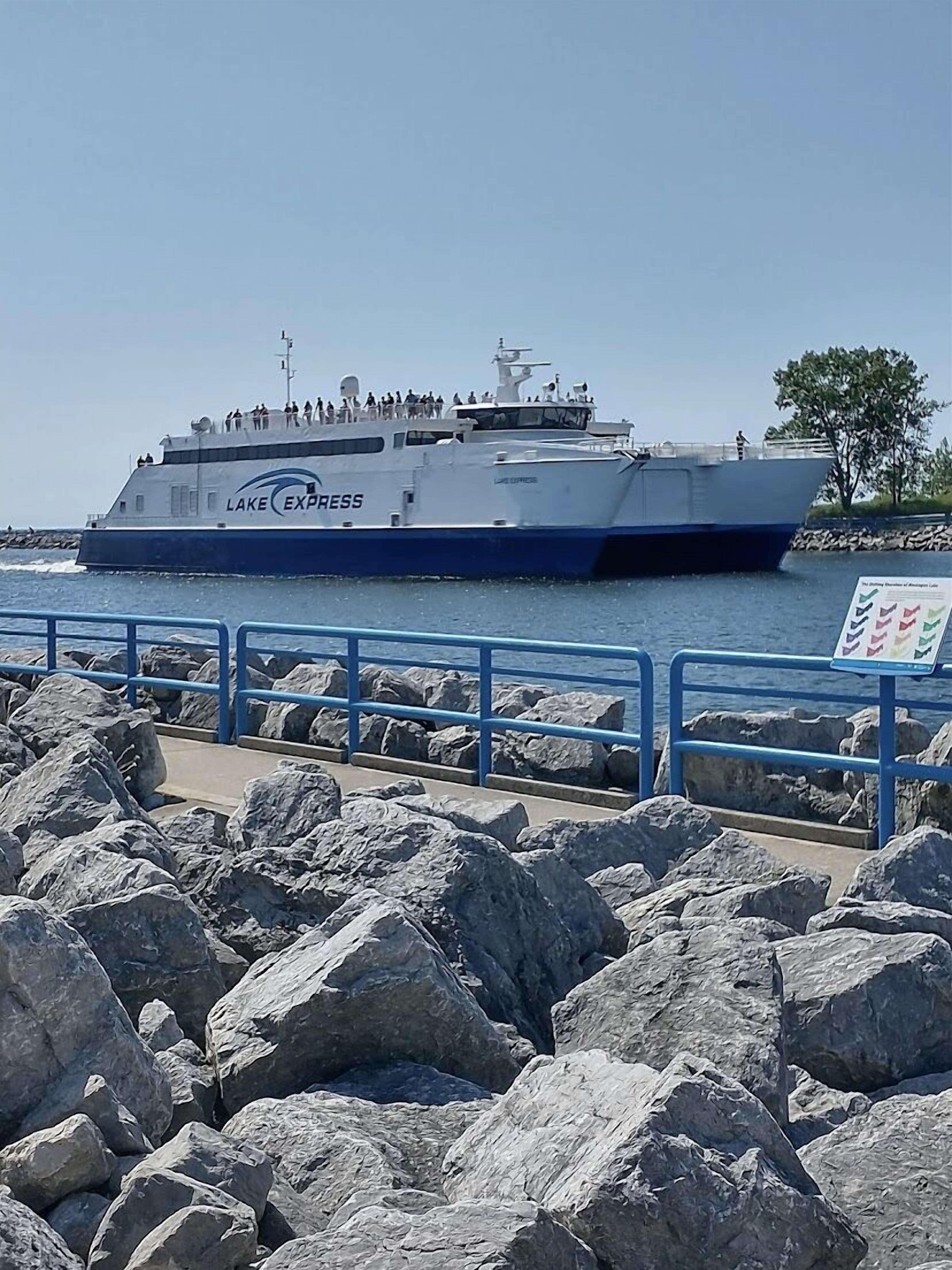 Een grote veerboot genaamd 'Lake Express' aanmeren bij een steiger met een rotsachtige kustlijn en heldere lucht.
