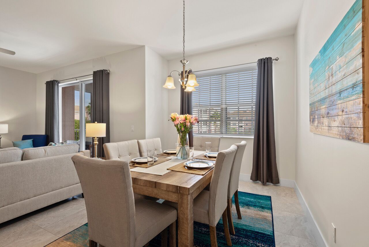 Un comedor con una mesa de madera dispuesta para cuatro, una araña y vistas al exterior a través de amplios ventanales con cortinas.