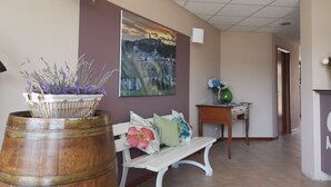 A hallway with a wooden barrel, a white bench, a painting of a landscape, and a small table with a vase. - Sportland (Colle di Val d'Elsa)