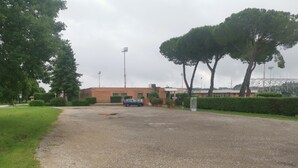 A gravel parking area with a blue car parked in front of a building with green windows and a brick facade. - Sportland (Colle di Val d'Elsa)
