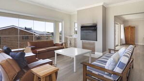 A spacious living room with a large window, a flat-screen TV, leather sofas, a wooden coffee table, and a wooden bench. - Ocean View Family Beach House (Hermanus)