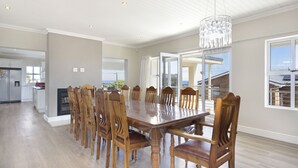 A spacious dining area with a large wooden table and chairs, a chandelier, and large windows offering views of the outside. - Ocean View Family Beach House (Hermanus)