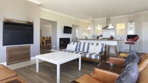 A modern living room with a white coffee table, striped sofa, and leather armchairs. A kitchen area with a bar and red bar stool is visible in the background. - Ocean View Family Beach House (Hermanus)