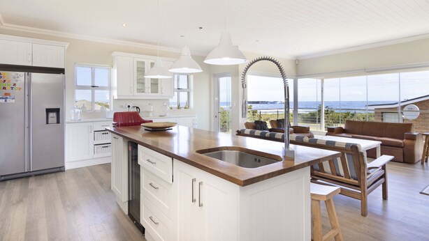 A modern kitchen with white cabinetry, a wooden countertop, and a large island with a sink. There is a refrigerator, a stove, and a window overlooking the ocean. - Ocean View Family Beach House (Hermanus)