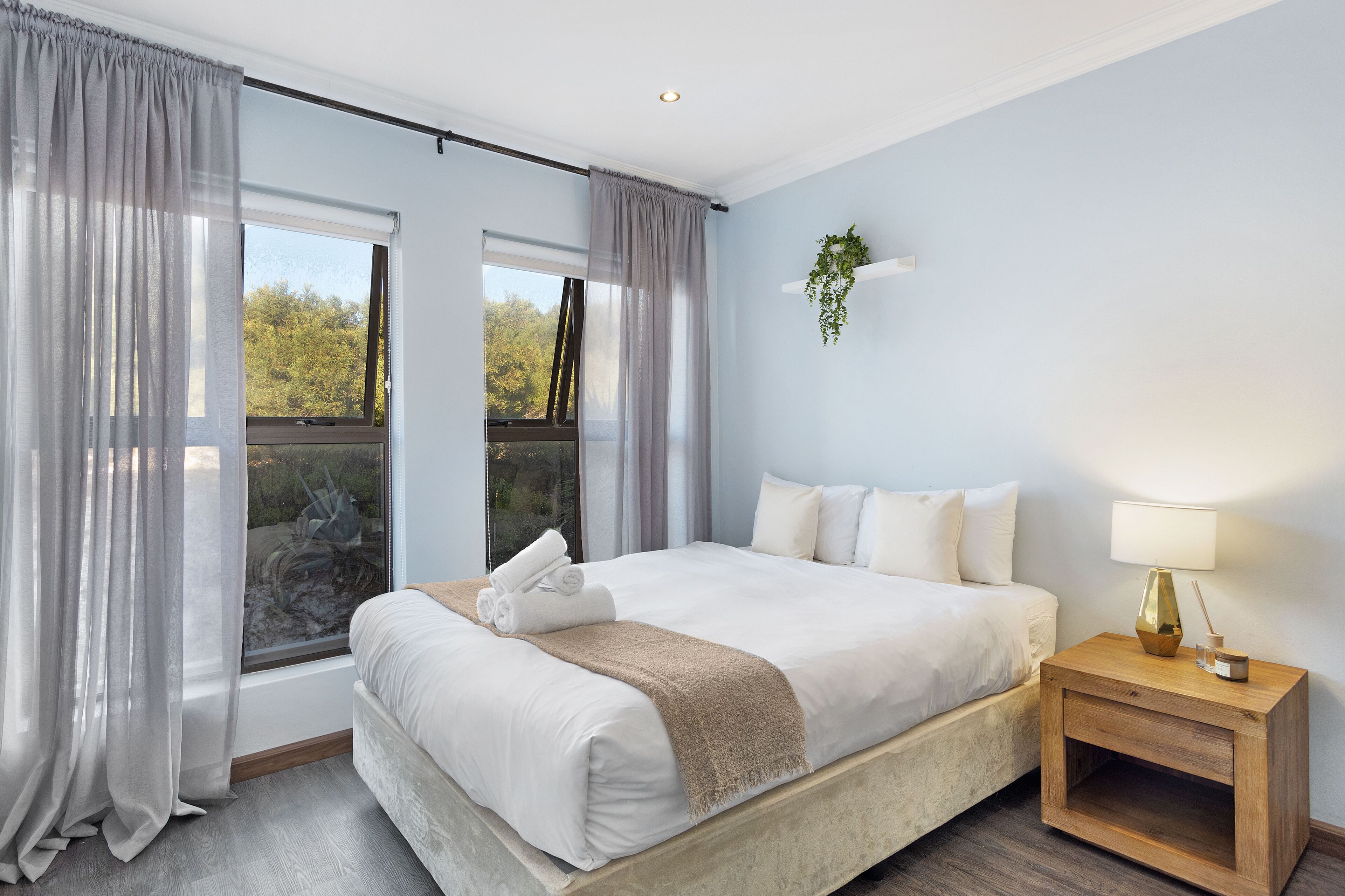 Un lit bien fait, avec une couette blanche et un plaid beige, une table de chevet avec une lampe, et une fenêtre ornée de rideaux légers.