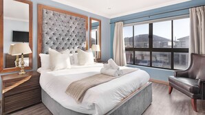 Quarto de hotel com uma cama grande, cabeceira estofada, janela com cortinas, poltrona de couro e mesa de cabeceira com luminárias.
