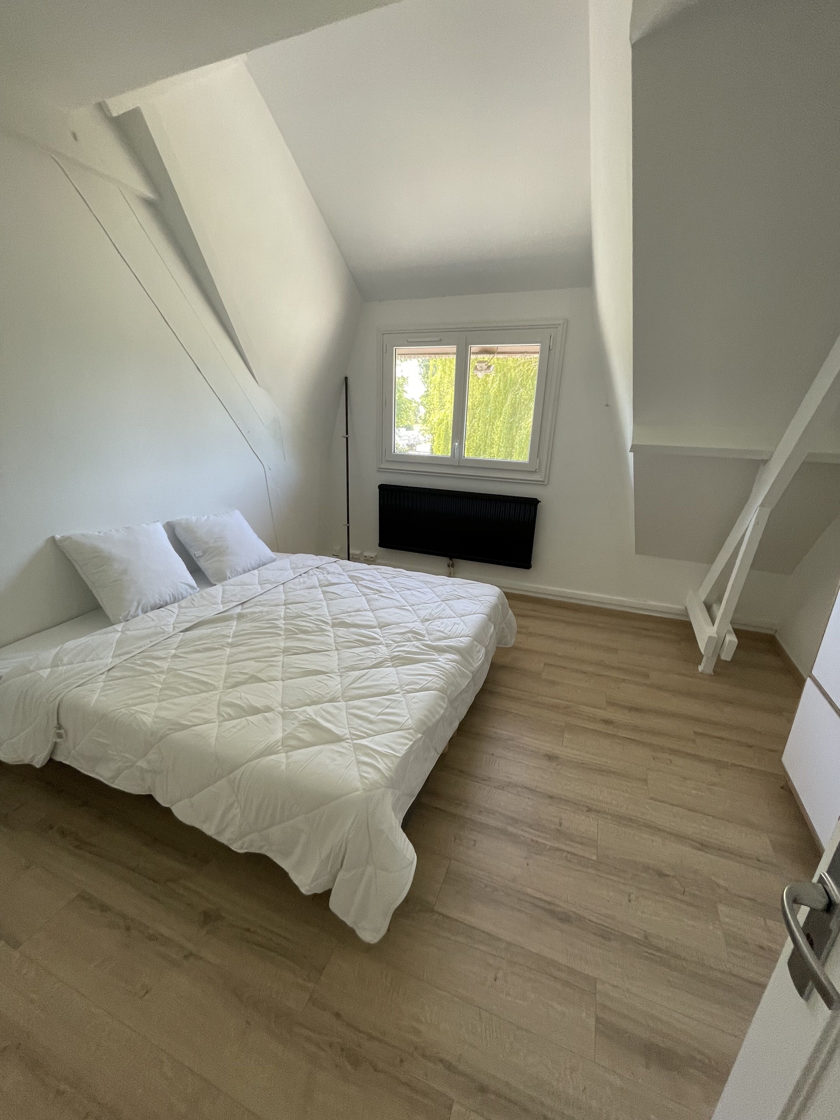Quarto com teto inclinado, cama com roupa de cama branca, radiador e janela com vista para áreas verdes.