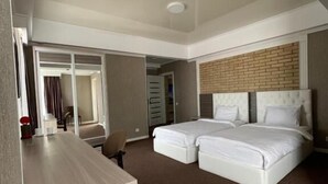 Quarto de hotel com escrivaninha, duas camas, parede decorativa de tijolos e vista para o corredor.