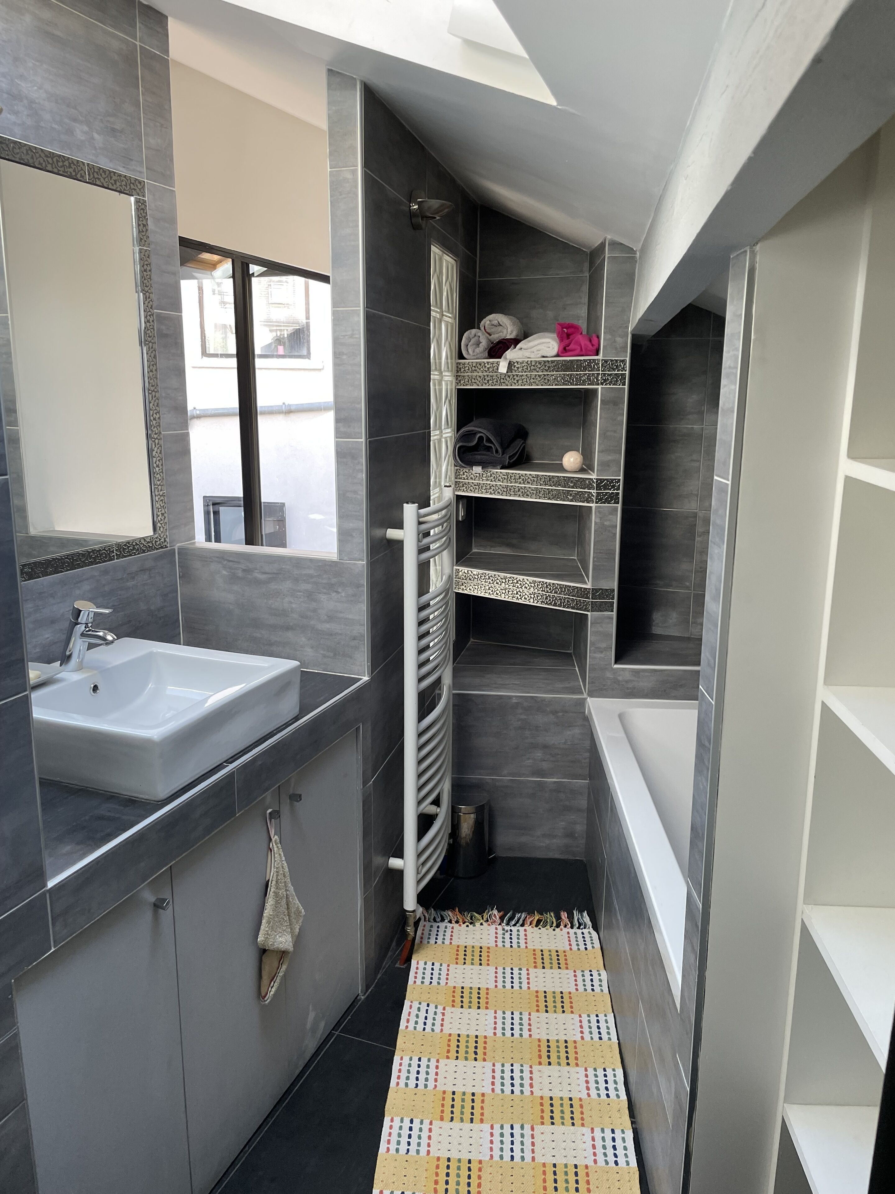 A modern bathroom with a white sink, dark tiles, and a patterned rug.