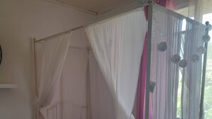 A bedroom with a four-poster canopy bed, a small bedside table with a vase of flowers, and a window with sheer curtains.