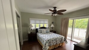 A bedroom with a bed, a ceiling fan, a window with blinds, a nightstand with a lamp, and a view of greenery outside. - Penthouse in Paradise! (Key West)