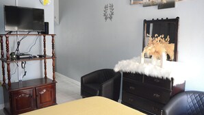 Une pièce comprenant un lit, un téléviseur posé sur un meuble, une commode, une chaise et une horloge murale.