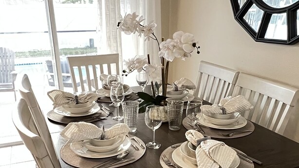 Une table à manger dressée pour six personnes, avec des chaises blanches, une nappe foncée et des fleurs d’orchidée blanches.