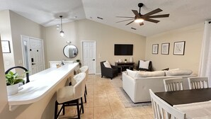 Une cuisine moderne avec un plan de travail blanc, un coin repas avec une table et des chaises, un salon avec un canapé et des fauteuils, et un ventilateur de plafond.