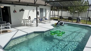 Une piscine avec des jeux gonflables, un patio couvert avec des chaises et une clôture blanche en arrière-plan.