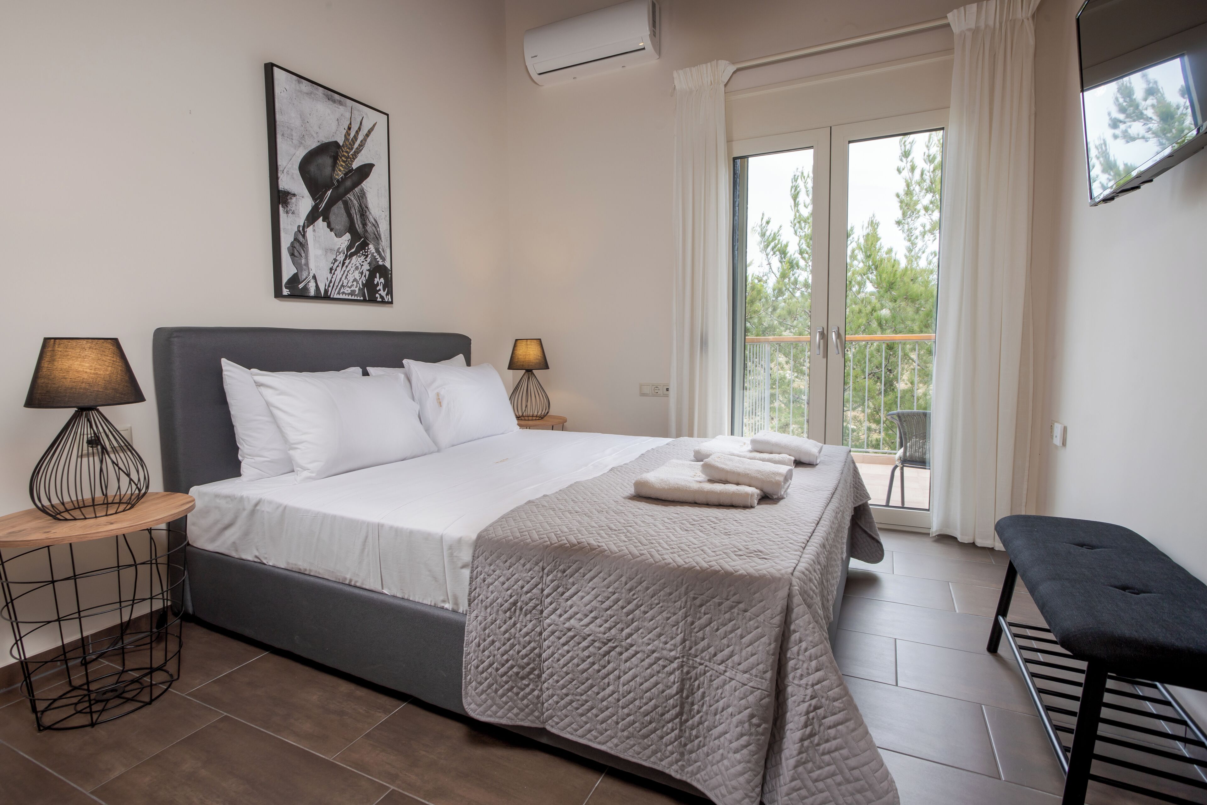 Un dormitorio con cama, dos lámparas de noche, un banco, un cuadro y un balcón con una silla.