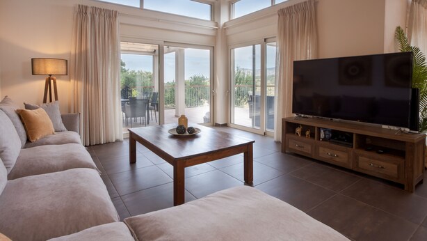 Un salon spacieux avec une grande télévision, un canapé, une table basse et un balcon agrémenté de rideaux.