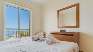 A bedroom with a bed, a wooden dresser, a mirror, and a view of the sea through a sliding door.