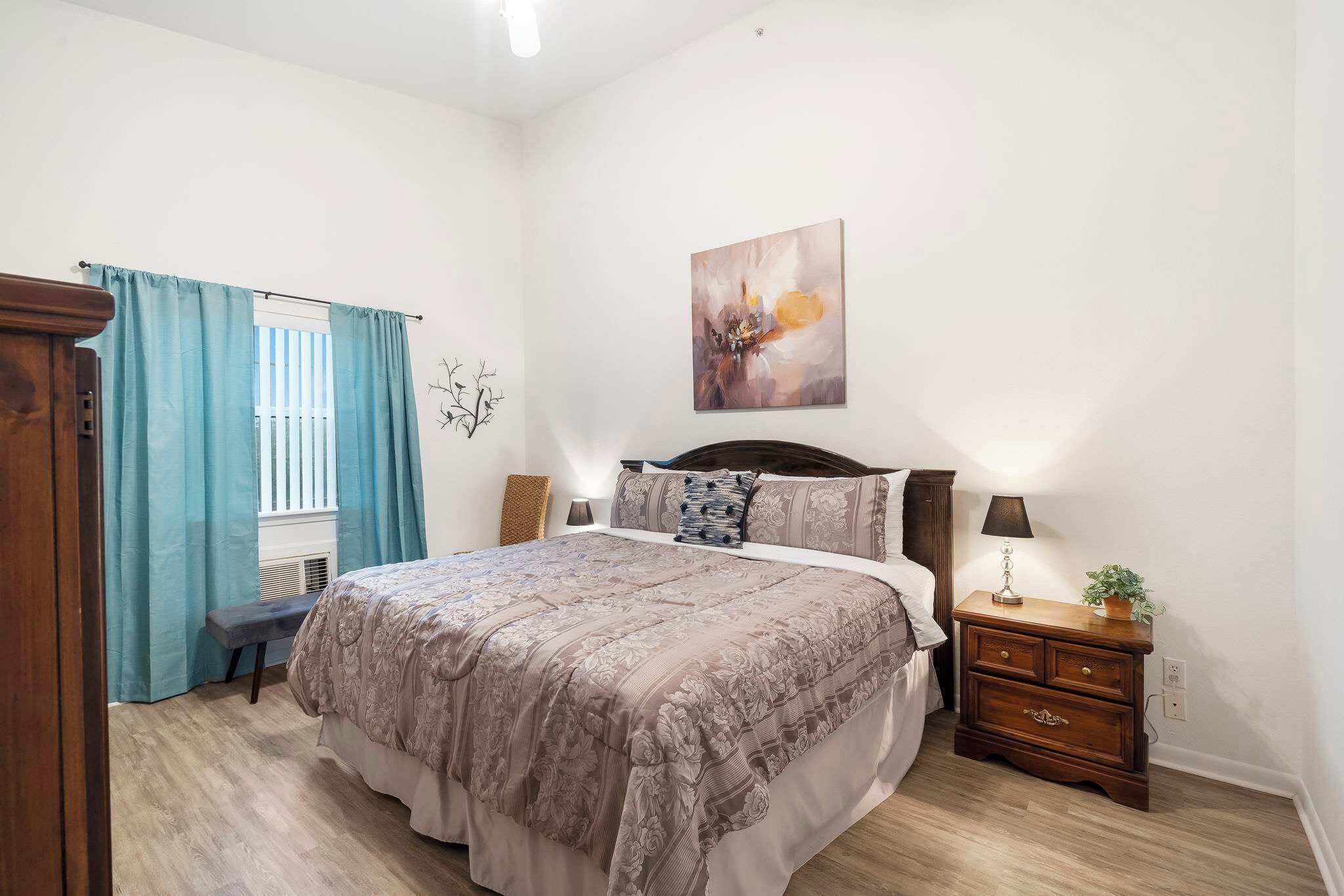 A bedroom with a large bed, a bedside table with a lamp, a wall-mounted artwork, and a window with curtains.