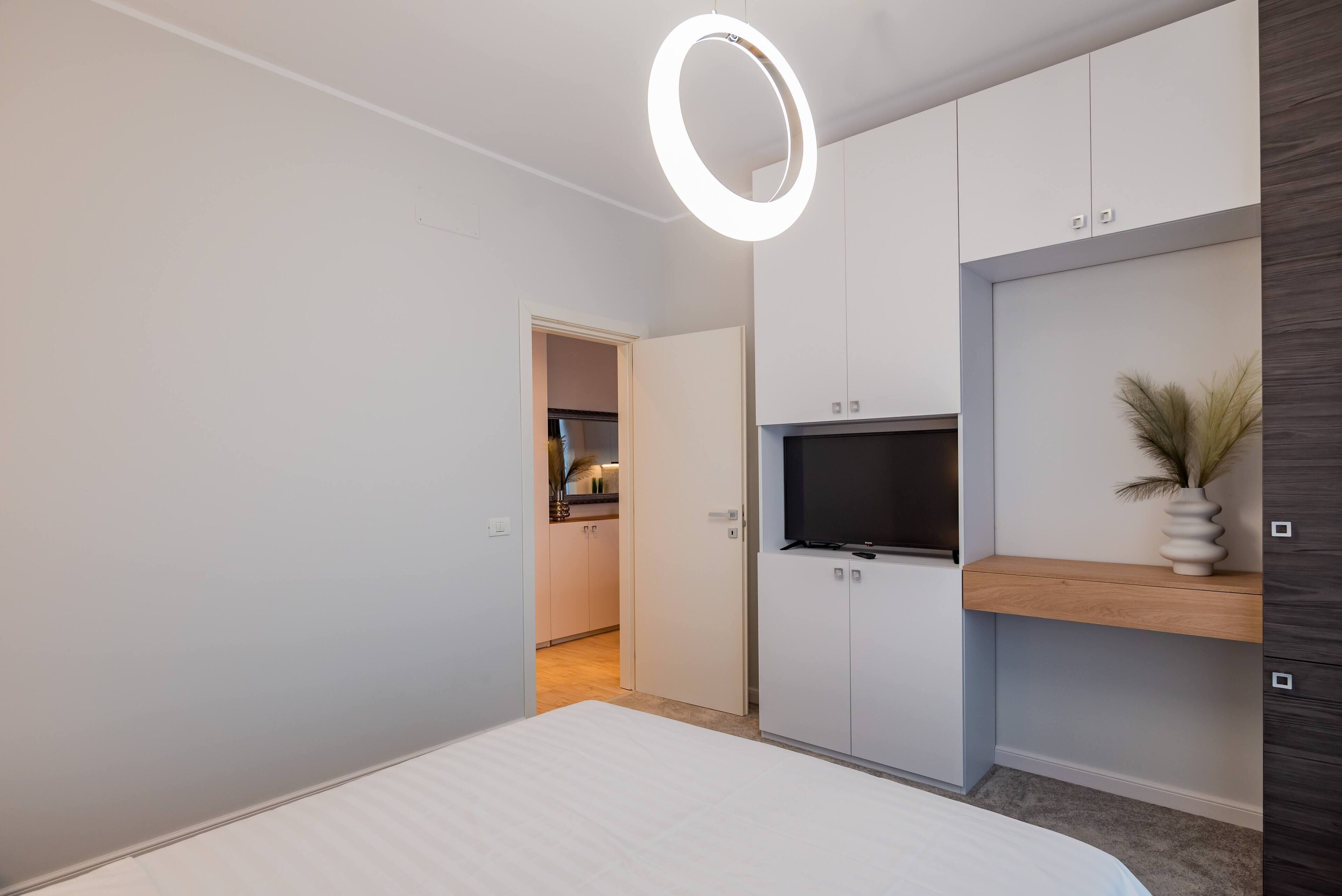 Ein modernes Schlafzimmer mit Bett, Fernseher und Kleiderschrank. Ein Flur führt zur Küche.
