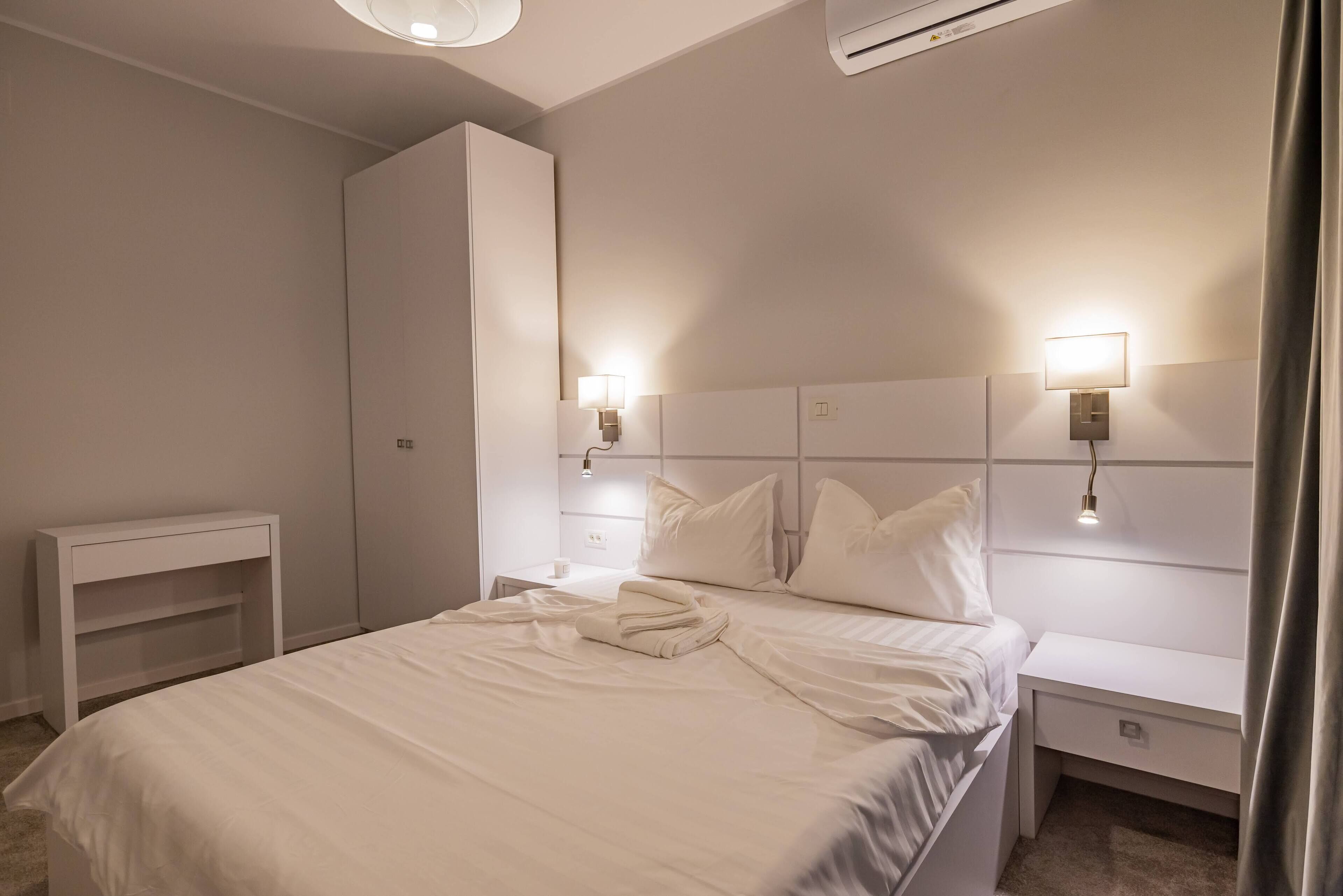 A modern hotel room with a neatly made bed, two bedside tables with lamps, a white wardrobe, and a small white console table.