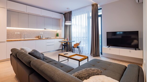 A modern living room with a sofa, a coffee table, and a TV. There is a kitchen area with cabinets and a dining table. - Ria Apartments (Navodari)