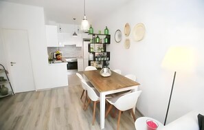 A modern kitchen with white cabinets, a wooden dining table, and white chairs. There is a small island with a sink and a fruit bowl.