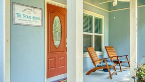 Un porche delantero con una puerta roja, dos sillas de madera con cojines azules y un cartel de bienvenida para The Pearl House en Amelia Island, Florida.