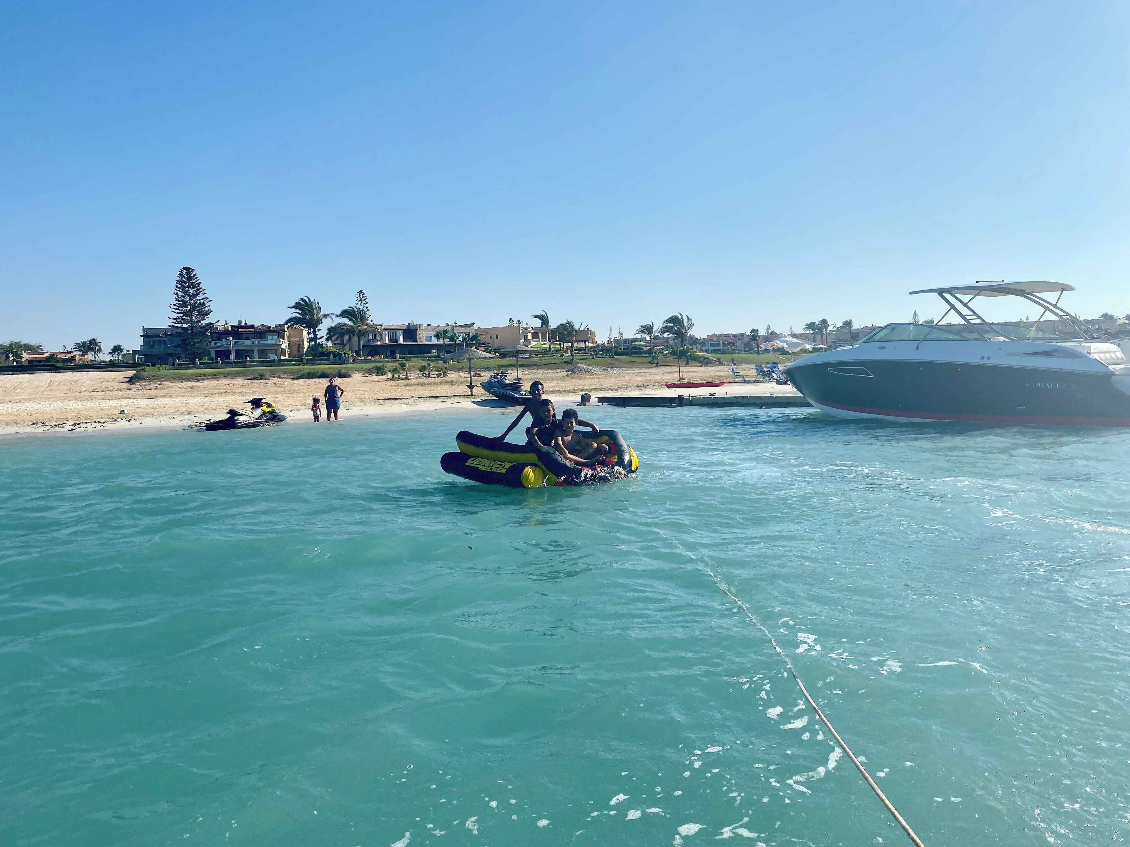 Une mer d’un bleu profond, avec un jet ski et un radeau gonflable sur l’eau. On aperçoit des bâtiments et une plage à l’arrière-plan.
