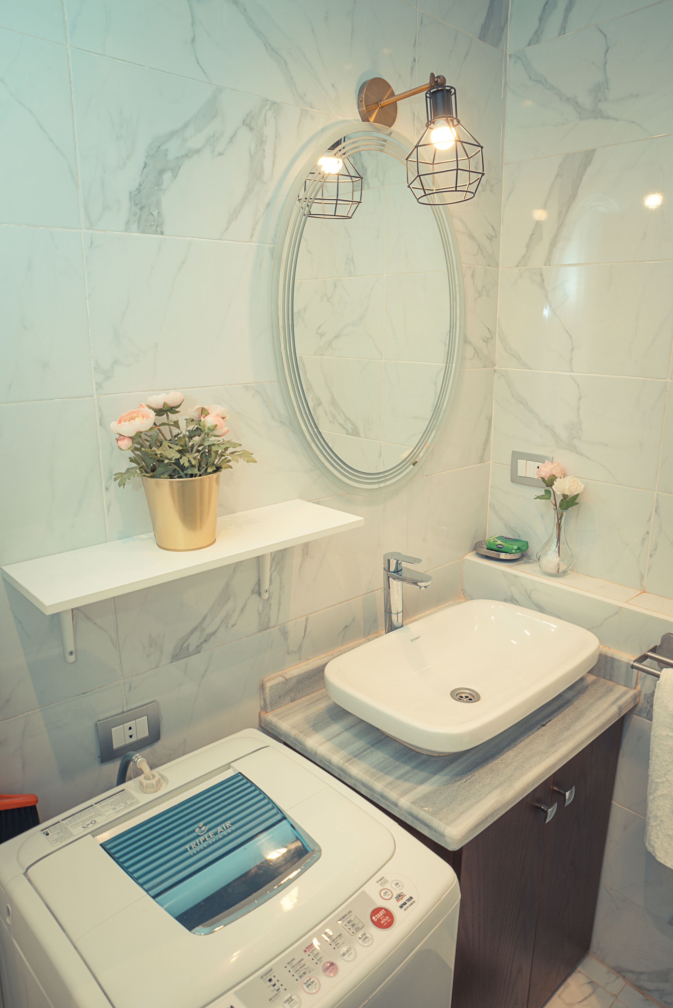 Un baño moderno con pared de mármol, un lavamanos independiente, una lavadora y un estante con una planta en maceta.